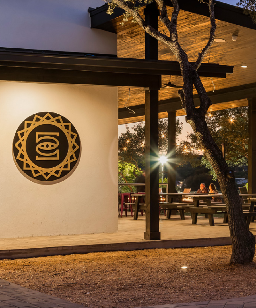 Outdoor evening view of the 5 Soul Wine Co. tasting area, featuring a large black and gold logo mounted on a white stucco wall. The warmly lit patio includes picnic tables where guests are enjoying wine under string lights, creating a cozy and inviting atmosphere surrounded by trees.
