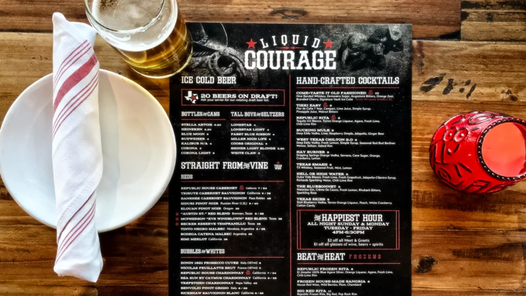 A rustic tabletop setting at The Republic Kitchen + Bar featuring a “Liquid Courage” drink menu listing beers, wines, and hand-crafted cocktails. The menu sits beside a white plate with a rolled napkin, a pint of beer, and a red glass candle holder on a wooden table.
