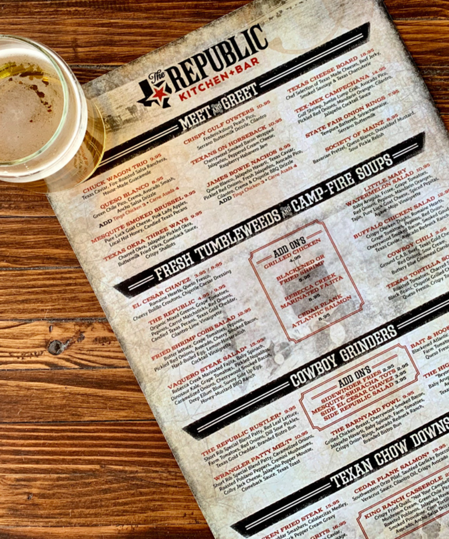 Food menu from The Republic Kitchen + Bar resting on a rustic wooden table beside a glass of beer. The menu sections include “Meet & Greet,” “Fresh Tumbleweeds,” “Camp-Fire Soups,” “Cowboy Grinders,” and “Texan Chow Downs,” featuring Southern-inspired dishes with bold typography and vintage design.