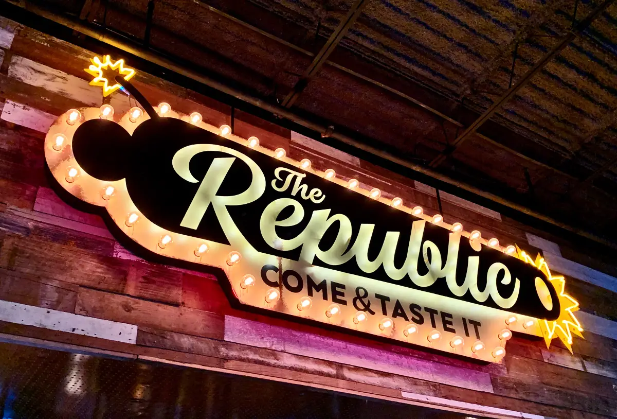 Illuminated marquee-style sign for The Republic Kitchen + Bar shaped like a cannon, with glowing bulbs around the border and neon accents. The sign reads “The Republic – Come & Taste It” and is mounted on a rustic wood-paneled wall inside the restaurant.
